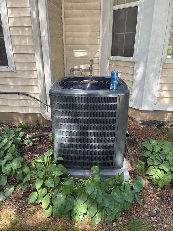 Completed installation of new Goodman air conditioning condensing unit on concrete pad against exterior wall. Unit properly positioned with adequate clearance for airflow, refrigerant lines and electrical conduit connected and secured, system tested and operational. Installation includes landscaping preservation around unit base with mulch bed maintained.