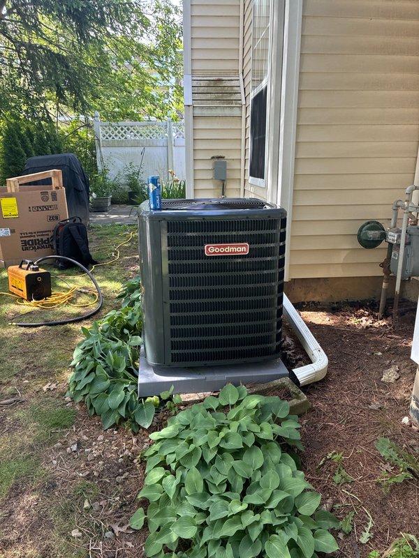 Completed installation of new Goodman air conditioning condensing unit on concrete pad against exterior wall. Unit properly positioned with adequate clearance for airflow, refrigerant lines and electrical conduit connected and secured, system tested and operational. Installation includes landscaping preservation around unit base with mulch bed maintained.