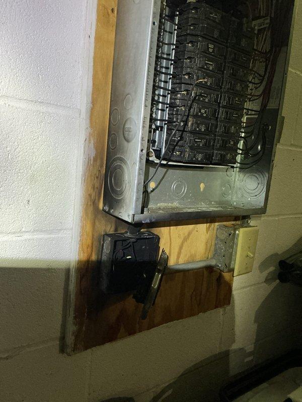 Responded to customer report of electrical box failure in kitchen wall. Upon inspection, documented existing electrical panel installation mounted on concrete block wall with visible water damage staining on wooden backing below panel. Panel contains multiple circuit breakers in older-style metal enclosure with gray finish. Photographed additional electrical components including black junction box and yellow connector box mounted on wooden backing, along with associated wiring. Also documented adjacent mechanical systems including water heater and plumbing infrastructure for comprehensive site assessment.