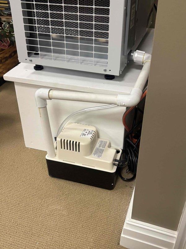Completed installation of dehumidifier system with condensate pump and drainage assembly for portable air conditioning unit. Installed 3/4" PVC piping and condensate tubing to properly route moisture removal from the dehumidifier through the condensate pump. Configured two-tier shelving system to accommodate both the air conditioning unit and condensate management equipment, ensuring proper drainage flow and operational efficiency.