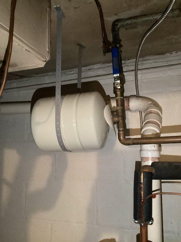 Performed plumbing inspection focusing on expansion tank system that has required multiple replacements in recent years. Examined horizontally-mounted white pressure tank with associated copper piping, PVC connections, brass fittings, and pressure gauge reading near bottom of scale. Documented condition of system components including visible corrosion and green patina on copper pipes, blue-handled valve, flexible corrugated hoses, and what appears to be a filter or pressure regulator in the assembly. Photographed installation for record purposes to track ongoing expansion tank performance issues.