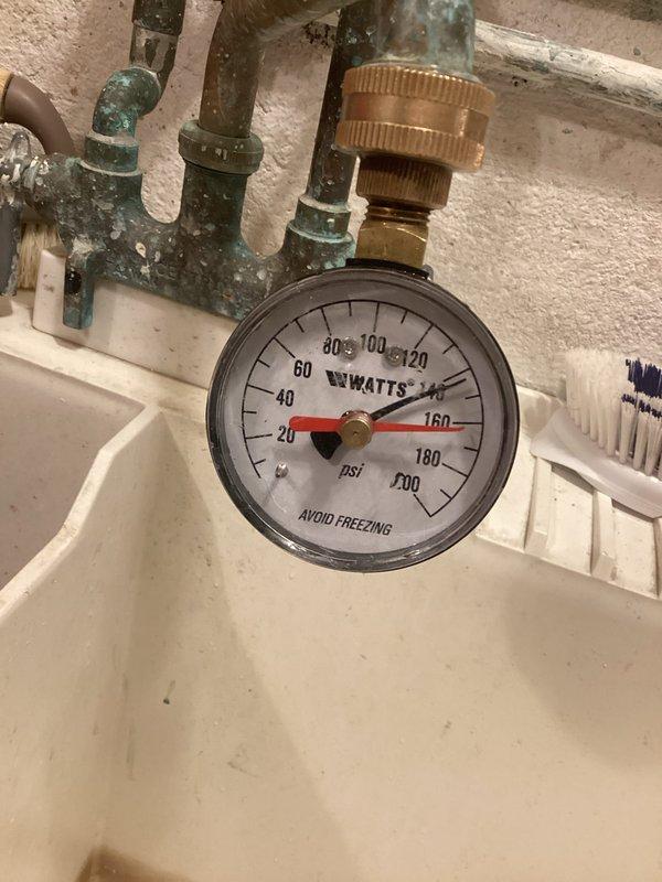 Performed plumbing inspection focusing on expansion tank system that has required multiple replacements in recent years. Examined horizontally-mounted white pressure tank with associated copper piping, PVC connections, brass fittings, and pressure gauge reading near bottom of scale. Documented condition of system components including visible corrosion and green patina on copper pipes, blue-handled valve, flexible corrugated hoses, and what appears to be a filter or pressure regulator in the assembly. Photographed installation for record purposes to track ongoing expansion tank performance issues.