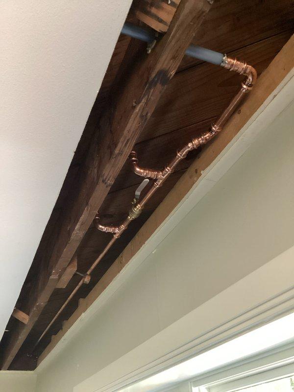 Returned to complete piping repair in exposed ceiling area with copper and PVC lines running along structural beams. Successfully finished installation of pipe joints, fittings, and valve connections, ensuring proper routing and secure mounting to ceiling joists.