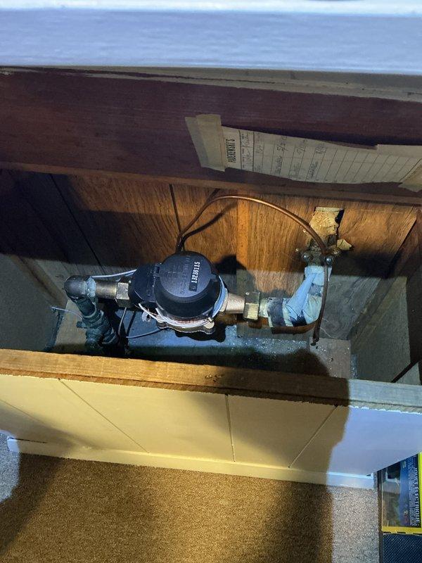 Completed mid-morning electrical inspection of residential property. Documented water meter installation in utility access area, including Neptune brand meter with copper and brass fittings connected to PVC piping, and photographed wall-mounted electrical control panel with multi-button interface in interior hallway for service records.