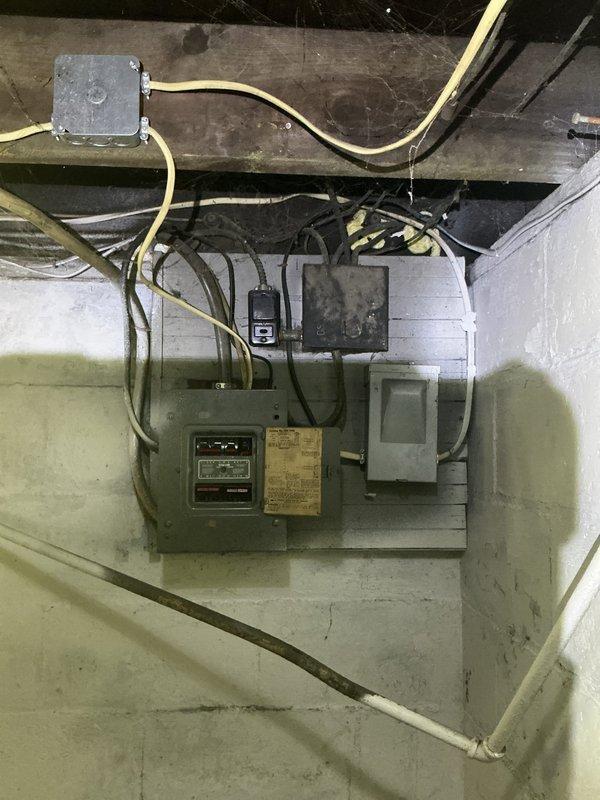 # Electrical Panel Assessment and Estimate

Conducted comprehensive evaluation of existing electrical panel and junction box system in basement utility area. Inspected aging panel board infrastructure including main electrical panel, auxiliary junction boxes, and associated wiring connections. Documented condition of gray-green main panel, secondary dark-colored electrical box with indicator display, and multiple conduit runs including cream-colored flexible conduit and black rigid conduit connections. 

Assessed internal wiring configurations within open junction box revealing brass and copper-toned connections and components. Evaluated mounting integrity on painted concrete block wall and examined condition of electrical connections running between panels and overhead junction boxes mounted to exposed floor joists. Photographed complete installation to document current state of electrical infrastructure for repair or replacement recommendation. Provided detailed estimate for panel bo
