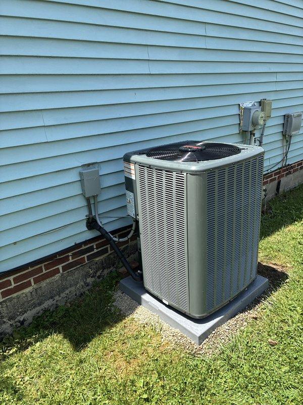 Completed installation of residential air conditioning condensing unit on concrete pad adjacent to building exterior. Unit properly positioned on level pad with electrical disconnect box mounted to siding, refrigerant line sets and electrical conduit professionally routed along exterior wall with appropriate insulation covering. DiversiTech line set cover installed to protect refrigerant lines, with PVC condensate drain line properly run to foundation area with secure fittings and foam insulation at bends.