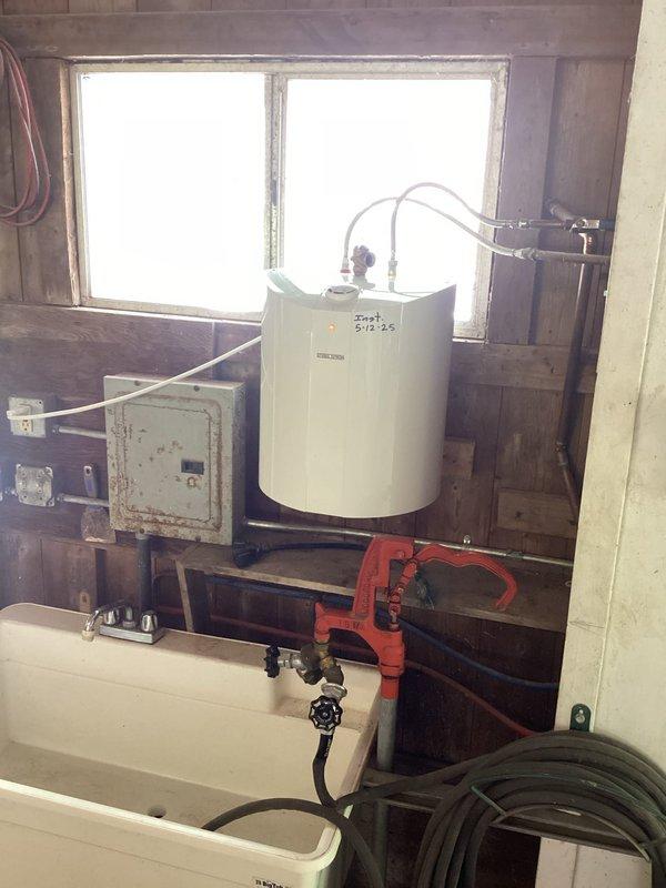Serviced small 5-gallon Stiebel Eltron electric water heater installed in barn utility space. Inspected unit mounted on wooden platform below window, verified electrical connections at adjacent panel, and examined plumbing connections including flexible supply hoses with brass valve assembly at top of tank. Confirmed temperature control dial operation and indicator light functionality to prepare unit for summer use.