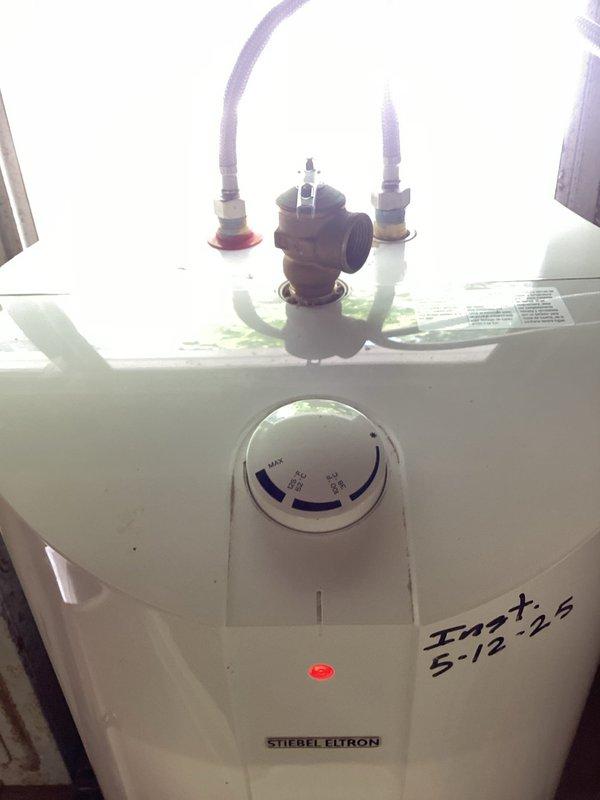 Serviced small 5-gallon Stiebel Eltron electric water heater installed in barn utility space. Inspected unit mounted on wooden platform below window, verified electrical connections at adjacent panel, and examined plumbing connections including flexible supply hoses with brass valve assembly at top of tank. Confirmed temperature control dial operation and indicator light functionality to prepare unit for summer use.