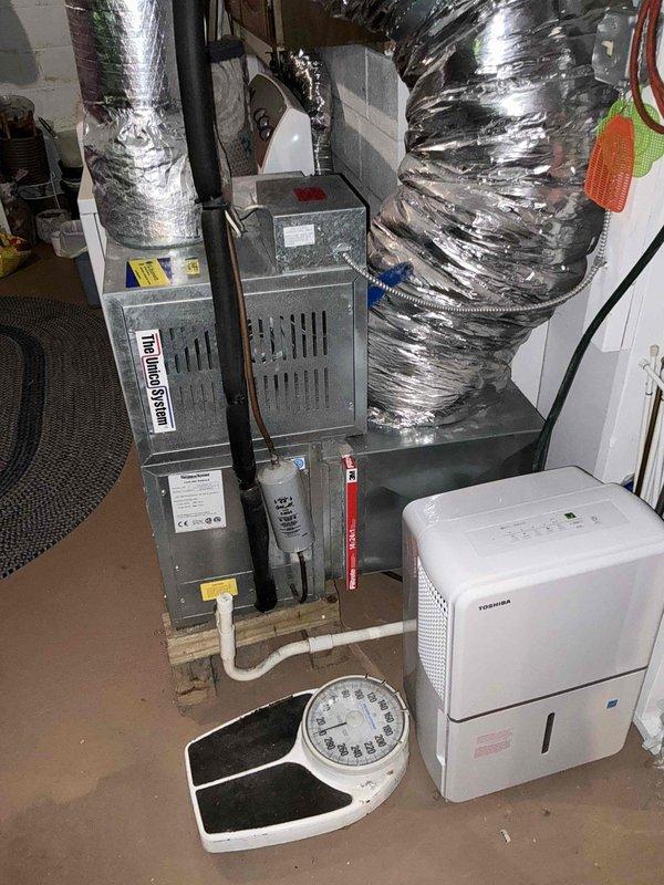 Conducted evaluation of existing 25-year-old HVAC system to provide replacement options. Inspected gray metal furnace unit with associated ductwork, piping connections, and ventilation components in basement utility area. Documented current equipment configuration including flexible ductwork routing and system layout to develop comprehensive replacement estimate for aging equipment.
