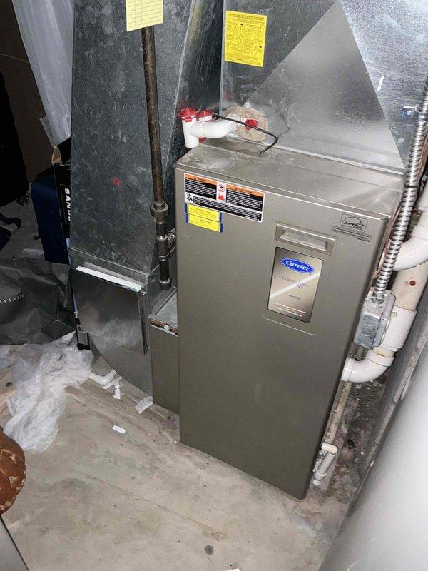 Provided comprehensive AC replacement options for existing Carrier HVAC system located in basement mechanical room with outdoor condenser unit positioned in landscaped area against building exterior. System currently features indoor air handler with ductwork connections and outdoor unit with refrigerant lines running through foundation, both units showing standard wear consistent with age. Equipment accessible for replacement with clear pathways for installation work.