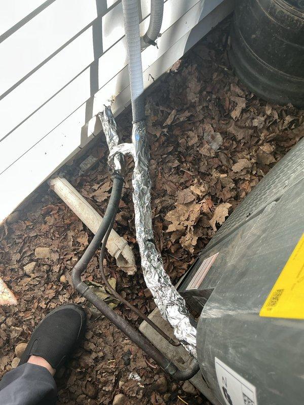 # HVAC Service Call - No Cooling Follow-Up

Responded to no cooling complaint on system that was not operating. This was a follow-up visit to a previous service call completed two weeks prior. Upon arrival, conducted visual inspection of outdoor condensing unit and associated refrigerant lines.

Exterior inspection revealed outdoor unit surrounded by significant leaf debris accumulation around the foundation area. Noted deteriorated insulation on refrigerant line set near building exterior, with yellow foam insulation showing signs of crumbling and damage. Line set insulation was compromised with exposed sections visible along the vertical run near the foundation.

Observed condensing unit location with multiple utility lines and cables running along ground level. Unit appeared accessible but surrounded by organic debris that could potentially restrict airflow or cause operational issues. Photographed equipment location and condition of refrigerant lines for documentation purposes.

Sy
