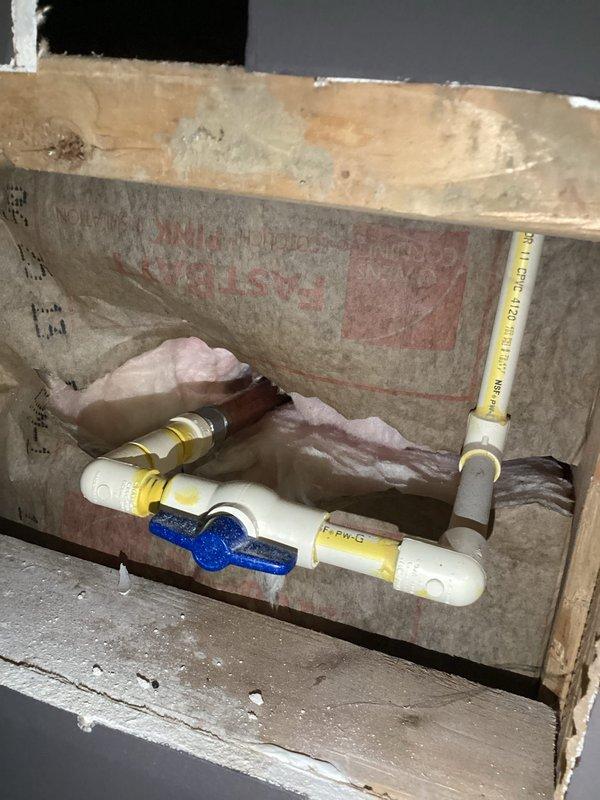 Responded to service call for exterior hose faucet requiring repair or replacement. Upon inspection, assessed the outdoor spigot connection mounted through vinyl siding and accessed the interior plumbing chase to examine water supply lines. Evaluated the brass fittings, PVC supply piping with blue shut-off valve, and connection points to determine necessary repairs and ensure proper functionality of the exterior hose bib system.