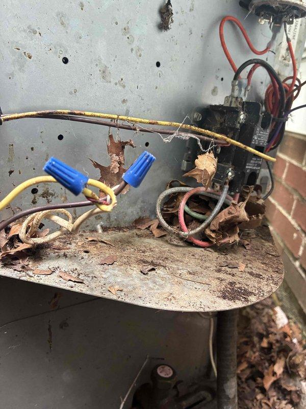 Responded to no cooling complaint and discovered severe electrical damage in outdoor unit control panel. Found extensive burn marks, charred wiring connections, and damaged wire insulation with exposed copper conductors throughout junction box. Blue wire connectors showed heat damage on yellow and brown wire connections. Heavy rust, corrosion, and organic debris accumulation indicated lack of maintenance and weather exposure. Verified electrical readings using digital multimeter. Documented all damaged components and hazardous conditions for repair recommendations.