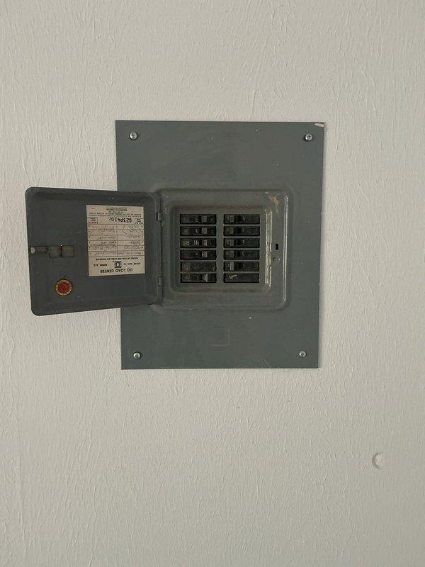 Conducted site assessment for exhaust fan installation estimate. Evaluated existing ventilation system mounted in utility space, documenting current blower assembly with light green metal housing and visible electrical wiring configuration. Inspected electrical service panel to verify available circuit capacity and breaker allocation for potential new exhaust fan installation. Photographed existing conditions including wall-mounted receptacles and ceiling texture to determine optimal mounting location and electrical routing requirements for proposed exhaust fan unit.