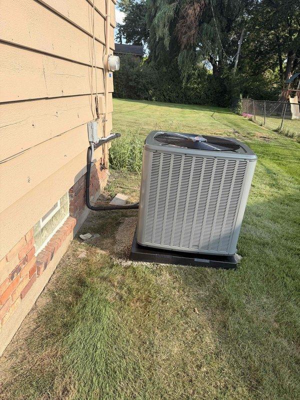Completed installation of new outdoor air conditioning unit on concrete pad adjacent to residential structure. Unit positioned on existing concrete foundation with proper clearances maintained from building exterior and landscaping. Electrical conduit run and connected from house to AC unit per code requirements. System tested and verified operational. Interior existing Rheem Criterion gas furnace remains in service with ductwork connections maintained for cooling distribution.