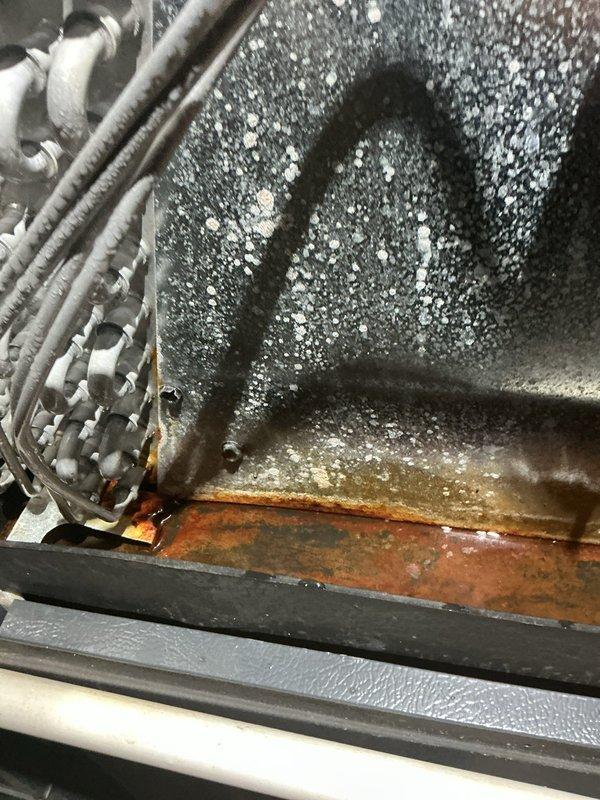 Responded to customer report of AC leak. Upon inspection, found evaporator coil completely frozen with heavy ice and frost buildup throughout the air handler unit. Observed significant rust and corrosion in the drain pan with standing water present, along with moisture accumulation on interior surfaces. The severe icing condition indicates restricted airflow or low refrigerant charge causing the coil to operate below freezing temperature. Documented conditions with photographs showing the extent of frost coverage on coil serpentine tubing and deteriorated drain pan requiring attention.