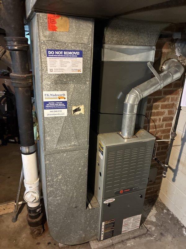 Completed installation of new air conditioning system including outdoor condensing unit and indoor air handler in basement mechanical room. Outdoor unit was positioned on concrete pad with proper clearances, refrigerant lines and electrical conduit professionally routed from exterior wall to equipment, and system installed on gravel bed adjacent to brick structure. Indoor air handler was mounted in basement with metallic ductwork connection, integrated with existing duct plenum system, and all required safety labels and manufacturer specifications properly affixed to equipment per code requirements.
