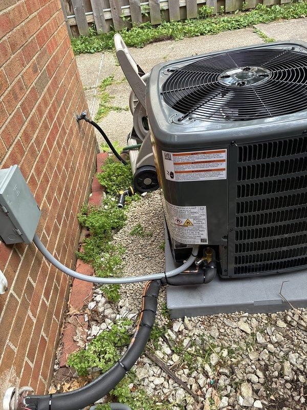 Completed installation of new air conditioning system including outdoor condensing unit and indoor air handler in basement mechanical room. Outdoor unit was positioned on concrete pad with proper clearances, refrigerant lines and electrical conduit professionally routed from exterior wall to equipment, and system installed on gravel bed adjacent to brick structure. Indoor air handler was mounted in basement with metallic ductwork connection, integrated with existing duct plenum system, and all required safety labels and manufacturer specifications properly affixed to equipment per code requirements.
