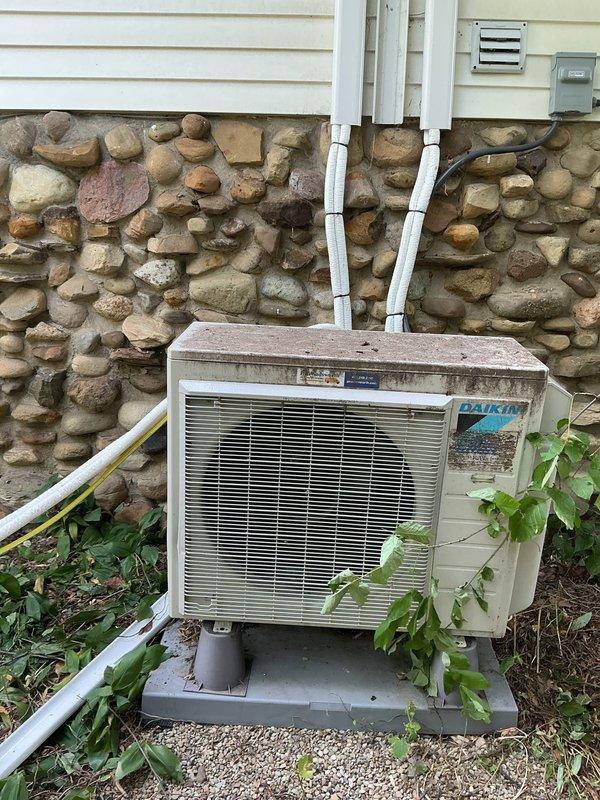 Responded to service call for Daikin split system requiring uninstallation and reinstallation due to roofing work in progress. Documented current installation condition of outdoor condenser unit mounted on composite pad against stone foundation with refrigerant lines and electrical connections running through exterior wall. Unit showed normal weathering and operational wear but no immediate mechanical concerns noted at time of inspection.