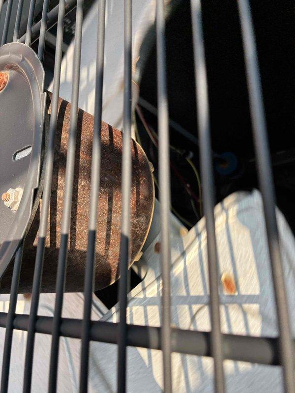 Responded to no cooling call with thermostat displaying "AC open circuit error" message. Upon inspection, found outdoor unit condenser coils with dust accumulation and compressor motor showing significant rust and corrosion on housing, indicating potential electrical or mechanical failure. Documented equipment condition through protective grille, noting deteriorated motor casing and corroded components that likely contributed to the open circuit fault detected by thermostat.