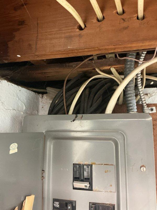 Conducted on-site assessment for electrical panel replacement in residential basement utility area. Evaluated existing gray metal panel mounted on concrete block wall, noting rust and discoloration around mounting points and reviewing current circuit breaker configuration. Documented existing electrical infrastructure including routing of various cables, conduits, and armored flex through ceiling joists, as well as proximity to plumbing systems and water heater for comprehensive replacement quote preparation.