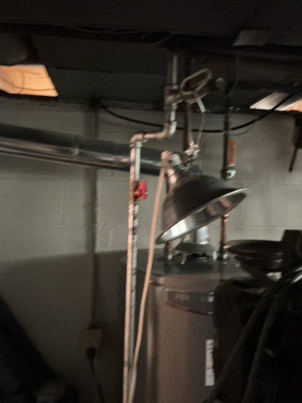 Conducted on-site assessment for electrical panel replacement in residential basement utility area. Evaluated existing gray metal panel mounted on concrete block wall, noting rust and discoloration around mounting points and reviewing current circuit breaker configuration. Documented existing electrical infrastructure including routing of various cables, conduits, and armored flex through ceiling joists, as well as proximity to plumbing systems and water heater for comprehensive replacement quote preparation.
