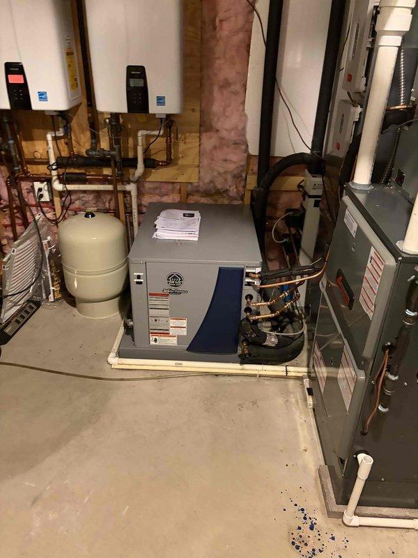 Completed geothermal HVAC system installation in residential mechanical room, including integration with existing infrastructure. System features modern geothermal heat pump unit with proper electrical specifications and safety labeling, coordinated with dual tankless water heaters, expansion tank, and air handler with refrigerant line connections. All equipment installed on protective floor pads with appropriate copper piping, PVC drainage, and electrical conduit routing per manufacturer specifications and code requirements.