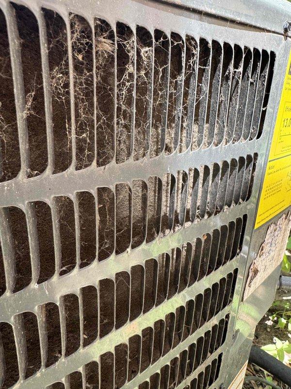 Responded to no cooling complaint on residential air conditioning system. Upon arrival, performed visual inspection of outdoor condensing unit. Found extensive cobweb accumulation across protective grille and significant debris buildup on condenser coil fins, restricting airflow. Condenser coil exhibited heavy layer of dust, dirt, lint, and fibrous material coating the metal fins and blocking spaces between fins. Documented conditions with photos showing weathered unit with poor maintenance history. Cleaning and restoration of proper airflow required to restore cooling capacity.