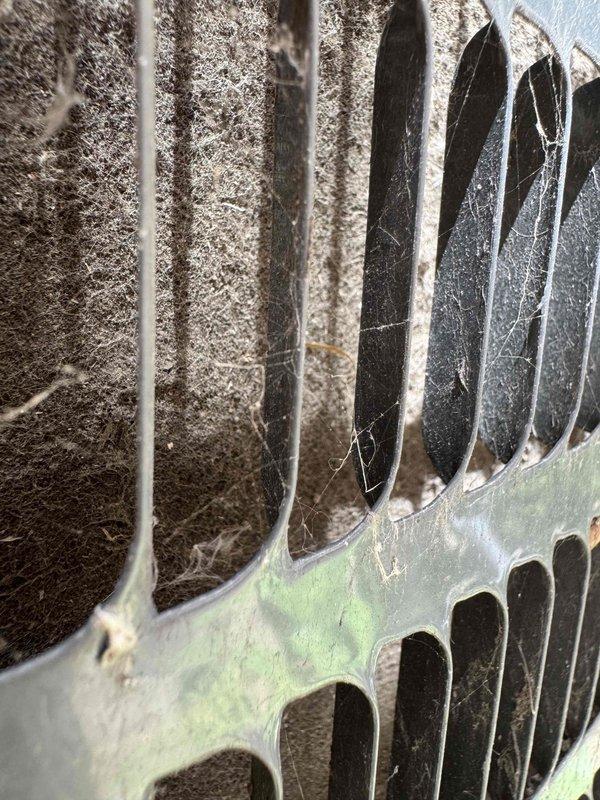 Responded to no cooling complaint on residential air conditioning system. Upon arrival, performed visual inspection of outdoor condensing unit. Found extensive cobweb accumulation across protective grille and significant debris buildup on condenser coil fins, restricting airflow. Condenser coil exhibited heavy layer of dust, dirt, lint, and fibrous material coating the metal fins and blocking spaces between fins. Documented conditions with photos showing weathered unit with poor maintenance history. Cleaning and restoration of proper airflow required to restore cooling capacity.
