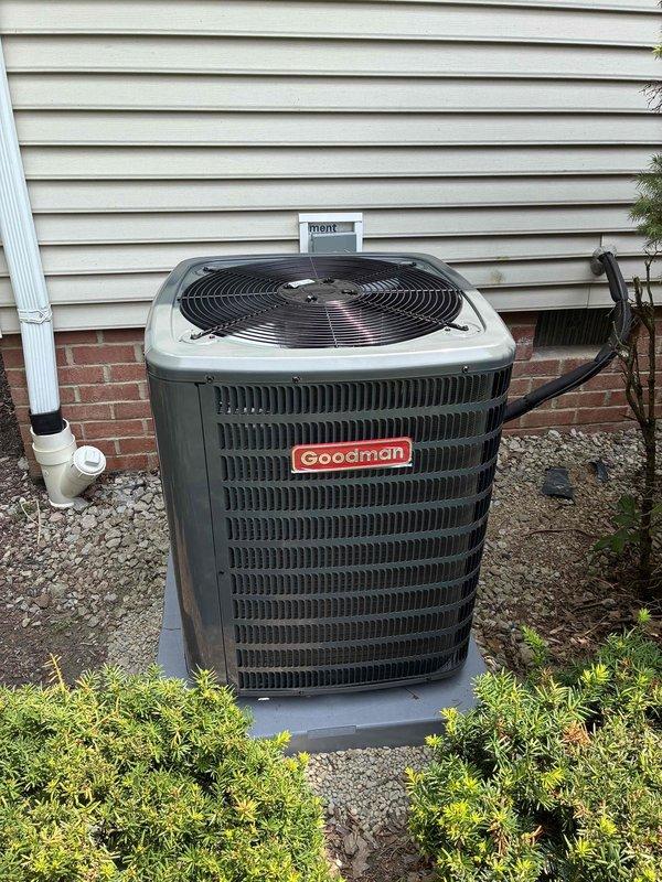 Completed installation of new Goodman condensing unit on concrete pad with proper gravel base and landscaping clearance. Unit positioned against exterior wall with all electrical conduit, disconnect box, and PVC condensate drain line properly installed and secured. System charged with refrigerant per manufacturer specifications, tested for proper operation, and verified all safety requirements met.