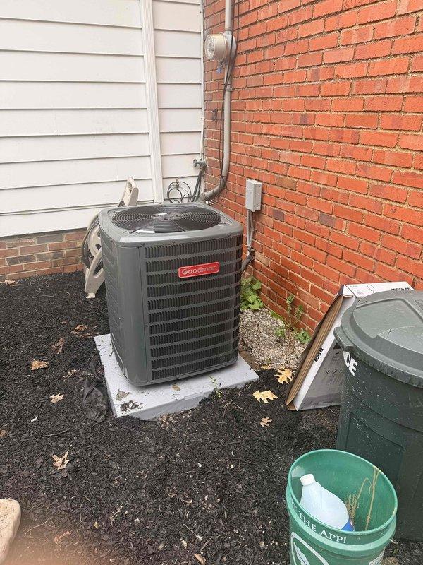 Completed installation of new Goodman air conditioning system including outdoor condensing unit positioned on concrete pad against building exterior. Installed metal conduit line set connecting outdoor unit to existing indoor air handler, with all electrical connections properly terminated and secured to building structure. System tested and confirmed operational upon completion.