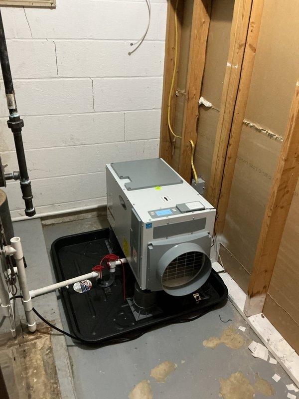 Completed HVAC estimate for whole home dehumidifier installation in basement utility area. Assessed existing mechanical space with white painted cinder block walls and concrete flooring for proper equipment placement and drainage requirements. Evaluated dehumidifier unit positioned on black condensate drain pan with appropriate clearances from exposed plumbing lines, wooden framing studs, and existing HVAC infrastructure. Verified control panel accessibility, circular duct outlet configuration, and condensate management system integration with existing PVC drainage piping. Documented installation location, spatial constraints, and mechanical connections required for proper indoor air quality system operation.