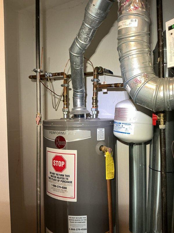 Responded to service call for excessive indoor humidity levels above 60% causing moisture damage to wooden flooring. Upon arrival, conducted thorough inspection of HVAC system and related equipment. Investigation revealed the issue was related to the water heater installation rather than the air conditioning system. Inspected Rheem Performance water heater and associated plumbing connections, including expansion tank, copper piping, and drain pan assembly. Documented equipment condition and installation setup with photographic evidence. Verified all safety labels and warning documentation were properly posted. Assessed concrete floor for water staining and examined drain pan for proper function to identify potential condensation sources affecting indoor humidity levels.