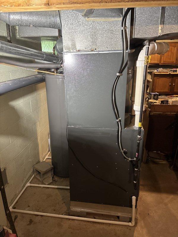Completed installation of residential HVAC furnace system in basement mechanical room. Installed gray metal tower-style air handler unit against concrete block wall, connected silver flexible ductwork to top of unit with proper metal strapping, and ran electrical conduit along right side of furnace secured with appropriate fasteners. Configured PVC condensate drainage piping and verified all connections for proper operation of the heating and cooling system.