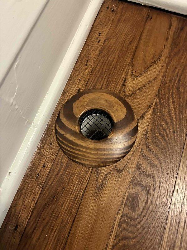 Completed installation of decorative wooden return air vent covers to match existing hardwood flooring and cabinetry. Installed floor register with concentric wooden ring design and central mesh grate, along with cabinet-mounted return air cover featuring natural wood grain finish and circular pull opening, ensuring proper airflow while maintaining aesthetic consistency throughout the space.