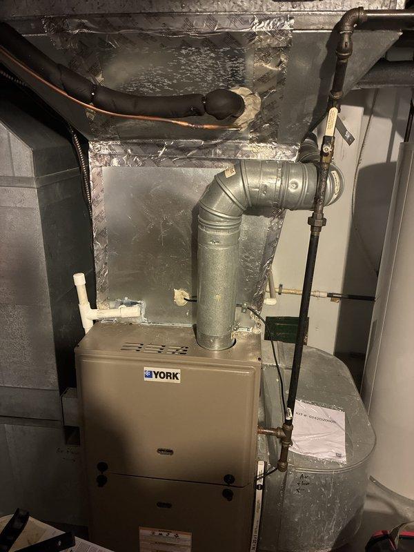 Completed full HVAC system installation including outdoor air conditioning unit positioned on concrete pad with proper electrical connections, junction box, and refrigerant lines secured to exterior wall. Installed York downflow furnace in basement utility area with galvanized metal ductwork and 90-degree elbow configuration, integrated with existing copper piping and gas lines. Whole home surge protection system installed and operational. Final cleanup and system commissioning completed following furnace installation, ensuring all components properly connected and tested for safe operation.