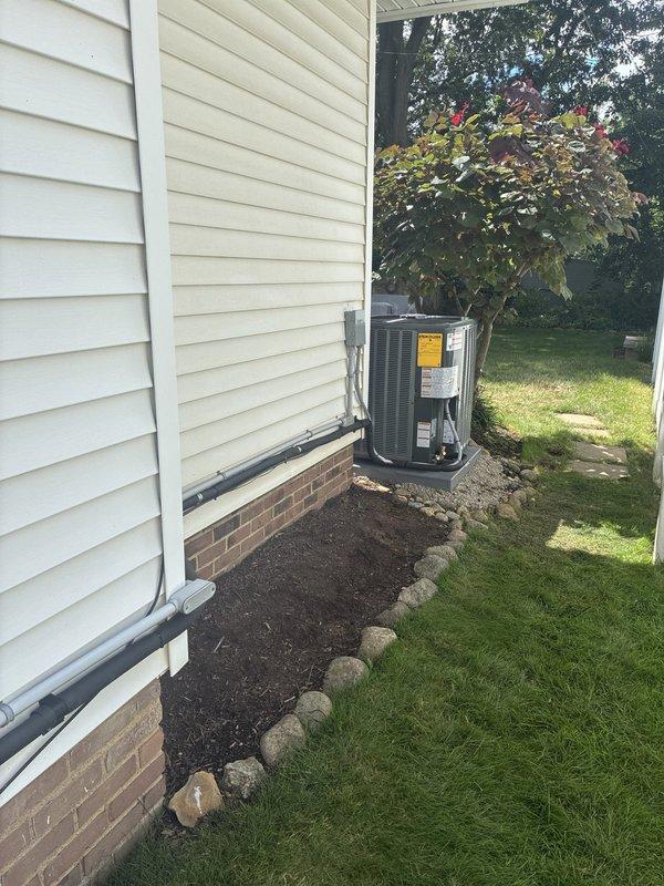 Completed installation of new residential air conditioning system at single-family home. Unit installed on concrete pad with gravel base and decorative rock border, positioned adjacent to house exterior with beige siding and brick foundation. Refrigerant lines routed and connected to indoor air handler, all electrical connections completed and tested. System startup performed, verified proper operation of compressor and condenser fan, confirmed adequate airflow and cooling capacity. Installation meets manufacturer specifications and local code requirements.