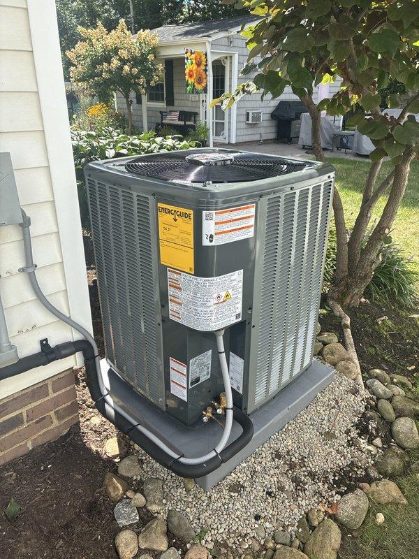 Completed installation of new residential air conditioning system at single-family home. Unit installed on concrete pad with gravel base and decorative rock border, positioned adjacent to house exterior with beige siding and brick foundation. Refrigerant lines routed and connected to indoor air handler, all electrical connections completed and tested. System startup performed, verified proper operation of compressor and condenser fan, confirmed adequate airflow and cooling capacity. Installation meets manufacturer specifications and local code requirements.
