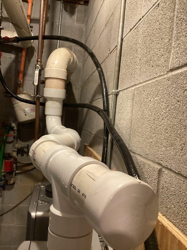 Completed installation of PVC elbows and intake screens on heating equipment in basement mechanical room. Documented existing HVAC system configuration including boiler unit with digital display and furnace with associated white PVC venting system. Obtained measurements of the installation area and captured detailed photographs of the mechanical space for combustion air calculation requirements. The work involved routing white PVC piping with multiple elbow joints along concrete block walls, ensuring proper venting configuration for the heating system. Verified all pipe connections and fittings were secure and properly aligned with existing equipment layout.