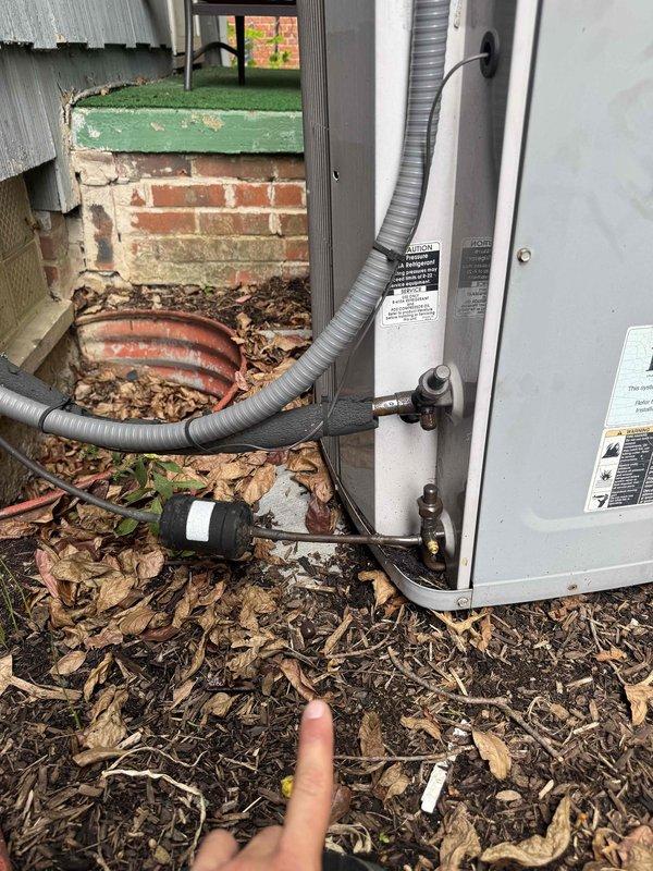 Responded to no cooling complaint on residential air conditioning system. Upon arrival, customer reported lack of cool air and suspected refrigerant leak. Performed visual inspection of outdoor condensing unit and observed signs of refrigerant loss at service valve connections. Identified active leak at brass valve fitting on refrigerant line near ground level, with visible oil residue and debris accumulation indicating prolonged seepage. Documented equipment condition with photographs showing weathered outdoor unit, exposed copper refrigerant lines, and leak point at valve connection. System requires refrigerant leak repair and recharge to restore cooling operation.