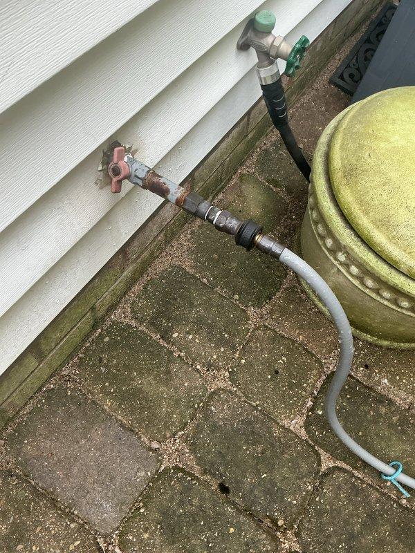 Responded to service call for non-functioning gas grill connection at residential property. Upon arrival, inspected exterior gas line assembly mounted on building foundation and confirmed customer report of connection issue at grill port. Found white flexible gas hose with adapter jammed into the connection point, preventing proper coupling with grill appliance. System featured copper gas piping with red-handled shutoff valve and secondary green-handled valve controlling gas flow to outdoor grill connection. Assessed corroded copper piping showing significant oxidation and deterioration, though still functional. Noted extensive moss and algae growth on foundation surfaces surrounding gas line installation, indicating prolonged moisture exposure in area. Yellow storage tank present adjacent to gas line assembly showed weathering consistent with outdoor exposure. Successfully freed jammed connection and verified proper gas flow to grill port, restoring functionality to outdoor cooking ap
