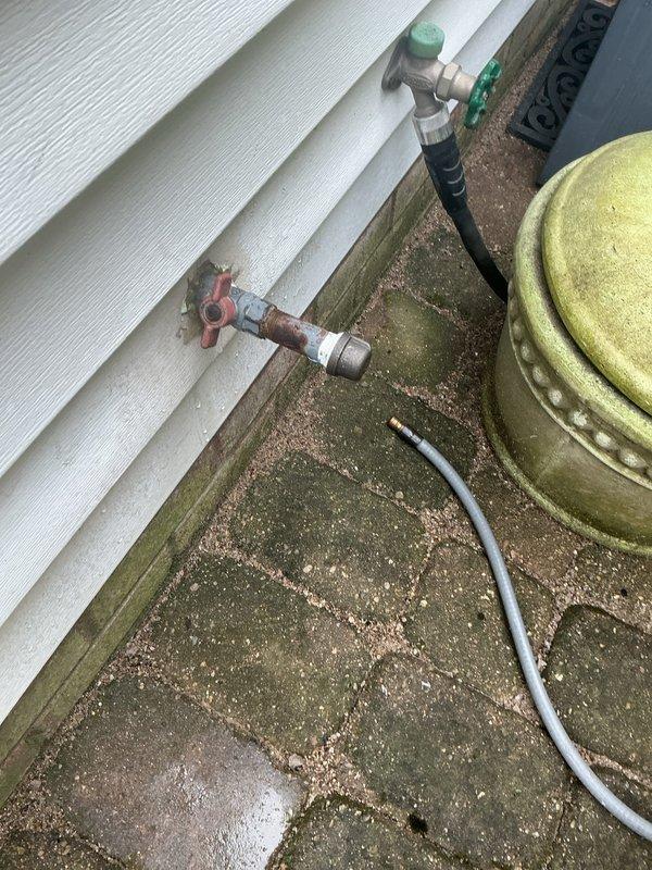 Responded to service call for non-functioning gas grill connection at residential property. Upon arrival, inspected exterior gas line assembly mounted on building foundation and confirmed customer report of connection issue at grill port. Found white flexible gas hose with adapter jammed into the connection point, preventing proper coupling with grill appliance. System featured copper gas piping with red-handled shutoff valve and secondary green-handled valve controlling gas flow to outdoor grill connection. Assessed corroded copper piping showing significant oxidation and deterioration, though still functional. Noted extensive moss and algae growth on foundation surfaces surrounding gas line installation, indicating prolonged moisture exposure in area. Yellow storage tank present adjacent to gas line assembly showed weathering consistent with outdoor exposure. Successfully freed jammed connection and verified proper gas flow to grill port, restoring functionality to outdoor cooking ap