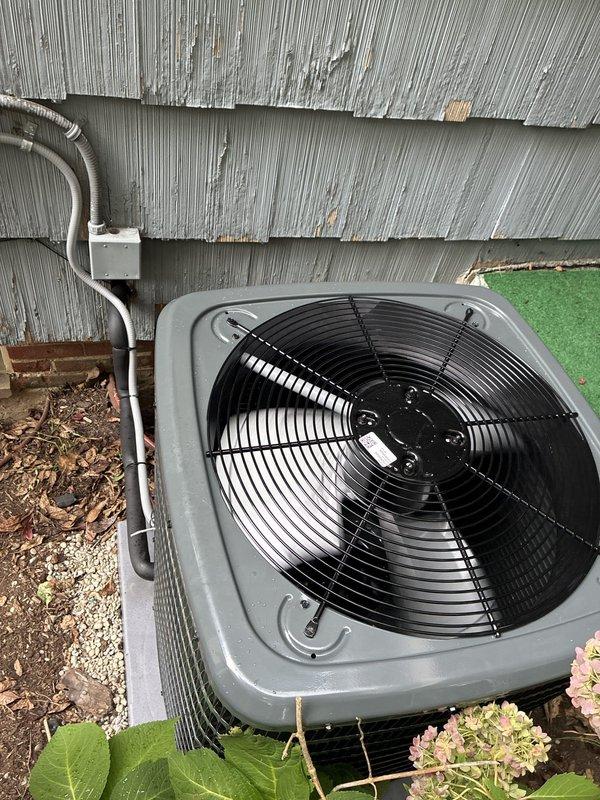Completed installation of new outdoor air conditioning condenser unit on concrete pad with gravel base. Installed copper refrigerant lines with insulation running from unit to building penetration point, secured white electrical conduit and junction box to exterior wall, and connected all electrical components. Unit positioned against building exterior with proper clearances, tested for operational functionality with fan assembly running correctly, and verified all connections secure. Installation area prepared with appropriate drainage and equipment properly leveled on pad.