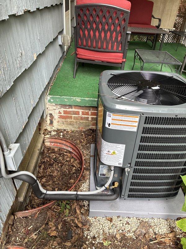 Completed installation of new outdoor air conditioning condenser unit on concrete pad with gravel base. Installed copper refrigerant lines with insulation running from unit to building penetration point, secured white electrical conduit and junction box to exterior wall, and connected all electrical components. Unit positioned against building exterior with proper clearances, tested for operational functionality with fan assembly running correctly, and verified all connections secure. Installation area prepared with appropriate drainage and equipment properly leveled on pad.