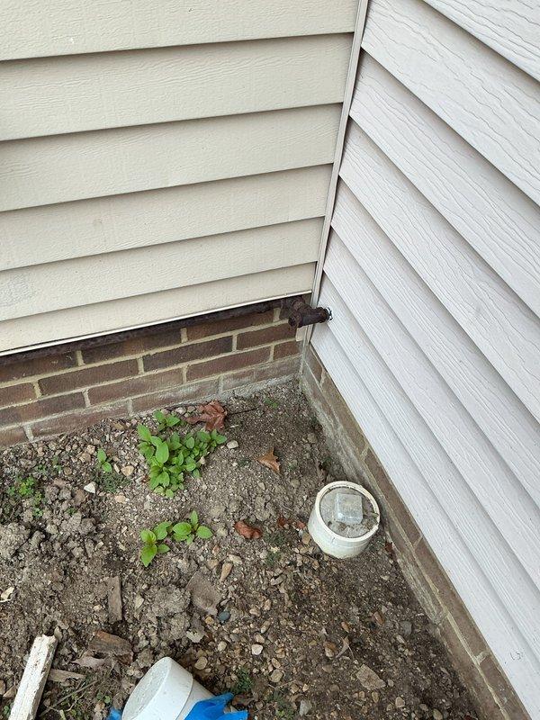 Conducted on-site assessment for gas line conversion project serving exterior fireplace. Existing black iron pipe currently runs approximately 25 feet along exterior foundation slab, requiring replacement with underground polyethylene gas line system. Evaluated current routing from interior connection point through basement area to exterior fireplace location. Documented existing black pipe configuration running exposed along building exterior where it is subject to weather exposure and potential damage. Assessed soil conditions at installation site, noting rocky, gravelly substrate with vegetation growth along foundation perimeter. Verified interior gas line connections and determined optimal transition point for new underground installation. Photographed existing plumbing infrastructure including drainage systems and structural elements to ensure proper coordination with proposed gas line burial. Recommended removal of existing exposed black iron piping and installation of approved p