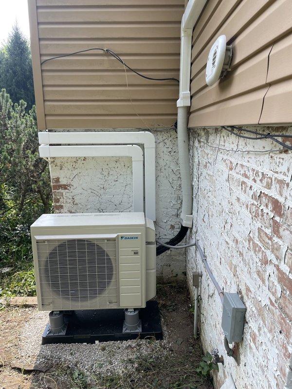 Completed installation of Daikin mini-split air conditioning system on residential property exterior. Mounted outdoor condenser unit on black pad at ground level with proper clearances, ran refrigerant line sets in white PVC covers vertically along building exterior to indoor unit location, and installed electrical conduit and wiring to power the system. All components secured to exterior wall surfaces, system tested and operational upon completion.