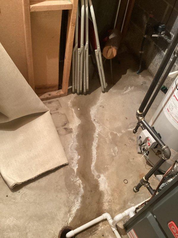 Responded to follow-up complaint regarding persistent moisture in basement utility area following recent pressure valve replacement. Upon inspection, observed significant water staining and white mineral deposits on concrete floor surrounding plumbing installation, indicating ongoing moisture infiltration despite dehumidifier operation. Examined pressure valve assembly mounted on concrete block wall, including red-handled control valve, connecting pipes, and transparent discharge tubing. Assessed water heater connections and associated plumbing components for potential leak sources. Documented floor conditions showing efflorescence patterns and discoloration consistent with continued water exposure. Verified valve installation integrity and investigated drainage system to identify source of persistent dampness in utility space.