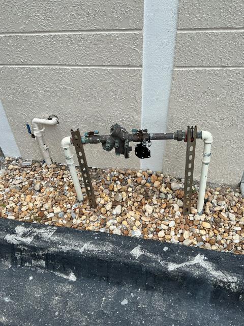 Plumbing system components including a backflow preventer and pipes on gravel surface, relevant to plumbing services in Boynton Beach, FL.