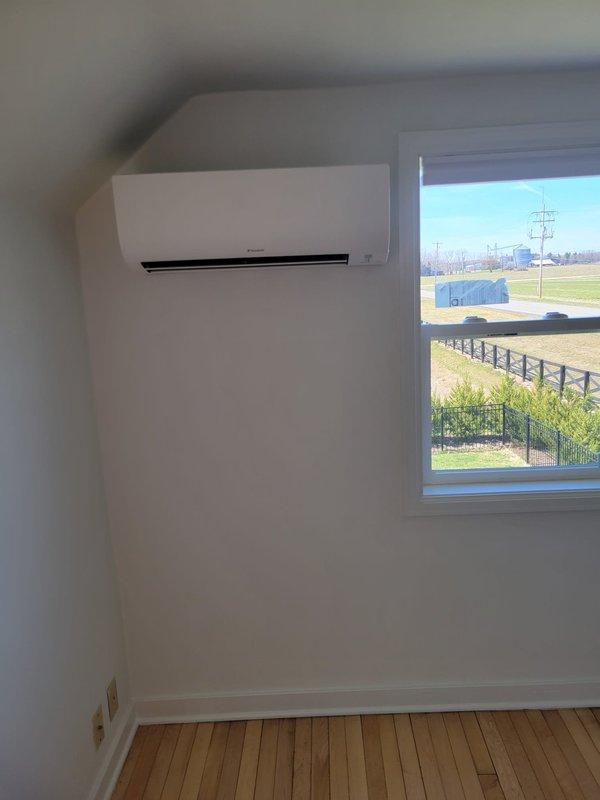 Completed full mini split system installation with indoor air handler mounted on interior wall to the left of window and outdoor condenser unit positioned on exterior with proper equipment pad and decorative rock bed for drainage. Ran refrigerant line set and electrical conduit from outdoor unit vertically along exterior siding to indoor unit, ensuring proper routing and professional appearance. System tested and operational upon completion.