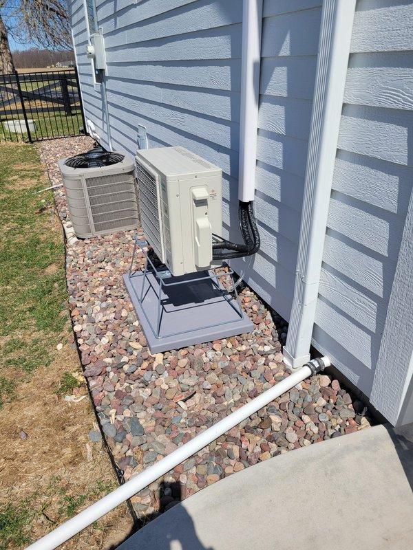 Completed full mini split system installation with indoor air handler mounted on interior wall to the left of window and outdoor condenser unit positioned on exterior with proper equipment pad and decorative rock bed for drainage. Ran refrigerant line set and electrical conduit from outdoor unit vertically along exterior siding to indoor unit, ensuring proper routing and professional appearance. System tested and operational upon completion.